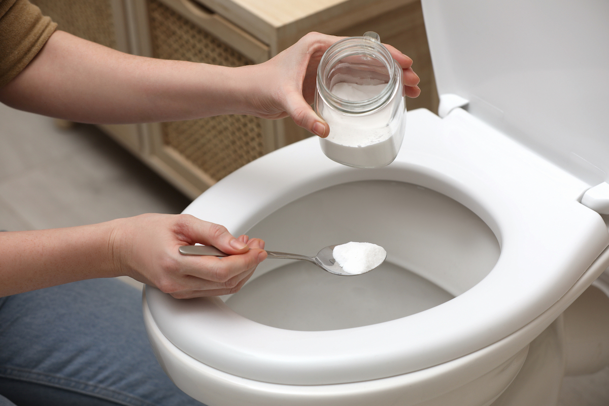 A person is scooping a spoonful of baking soda from a glass jar to put inside of a toilet.