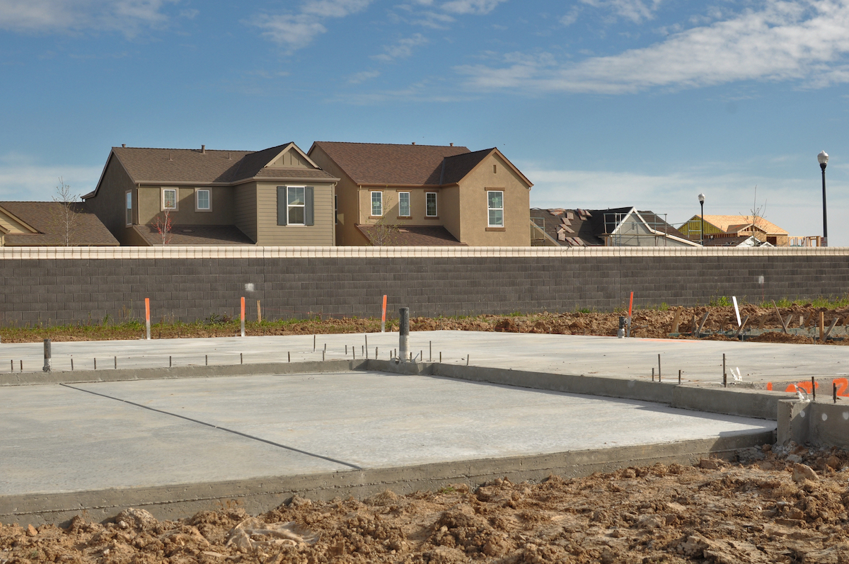 A new home's concrete slab foundation is laid in a dirt lot in front of a neighborhood.