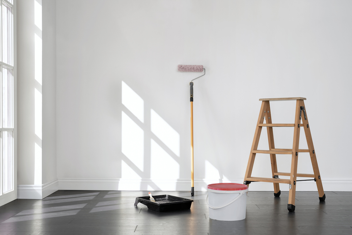 An empty room contains from left to right: a roller tray, a long-handled roller brush, a paint pail, and a step ladder.