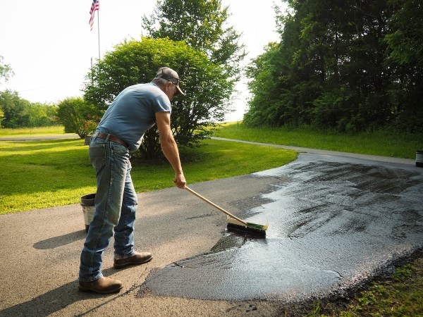 Older man in jeans and cap coats asphalt driveway with a seal using proper equipment.