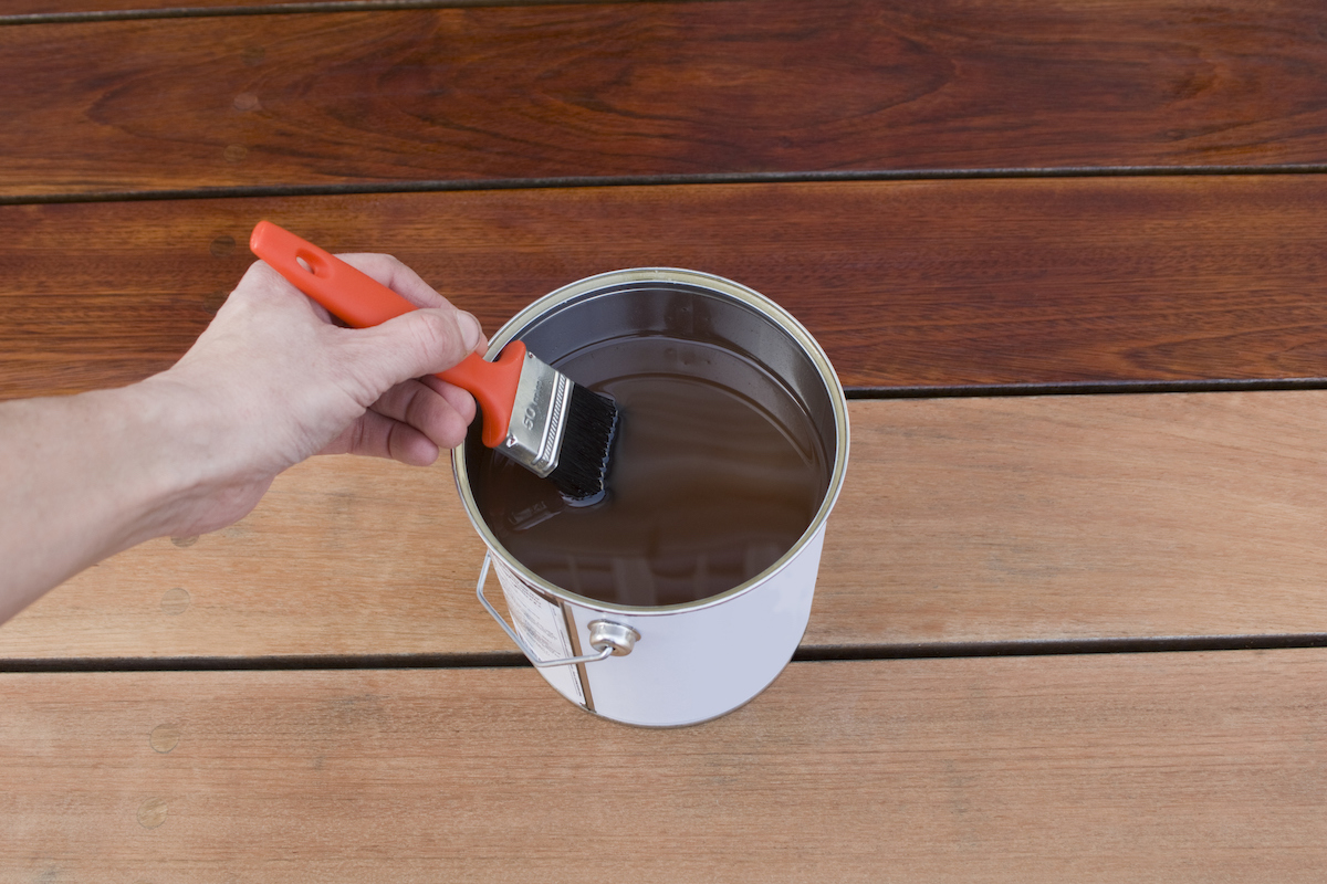 Person dips a paintbrush into a can of deck stain, with some stained and unstained boards underneath.
