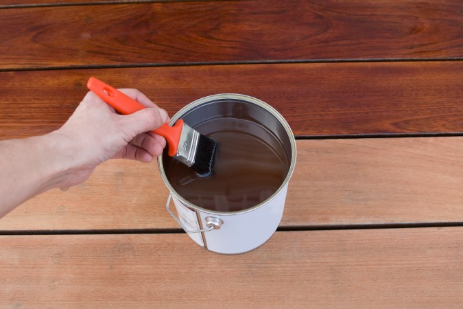 Person dips a paintbrush into a can of deck stain, with some stained and unstained boards underneath.