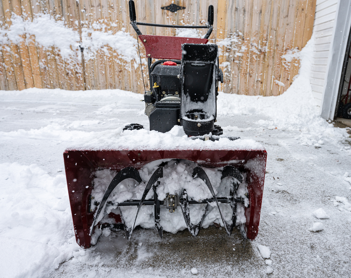 A snow blower with clogged blades sitting in a snowy driveway.