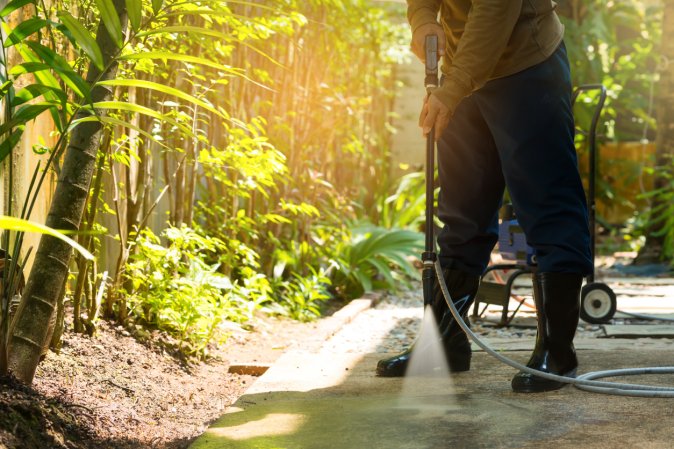 A person wearing rain boots pressure washing a concrete driveway.