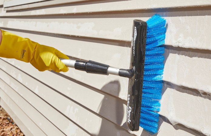 Woman with yellow rubber gloves uses a long-handled brush to clean beige vinyl siding on a home.