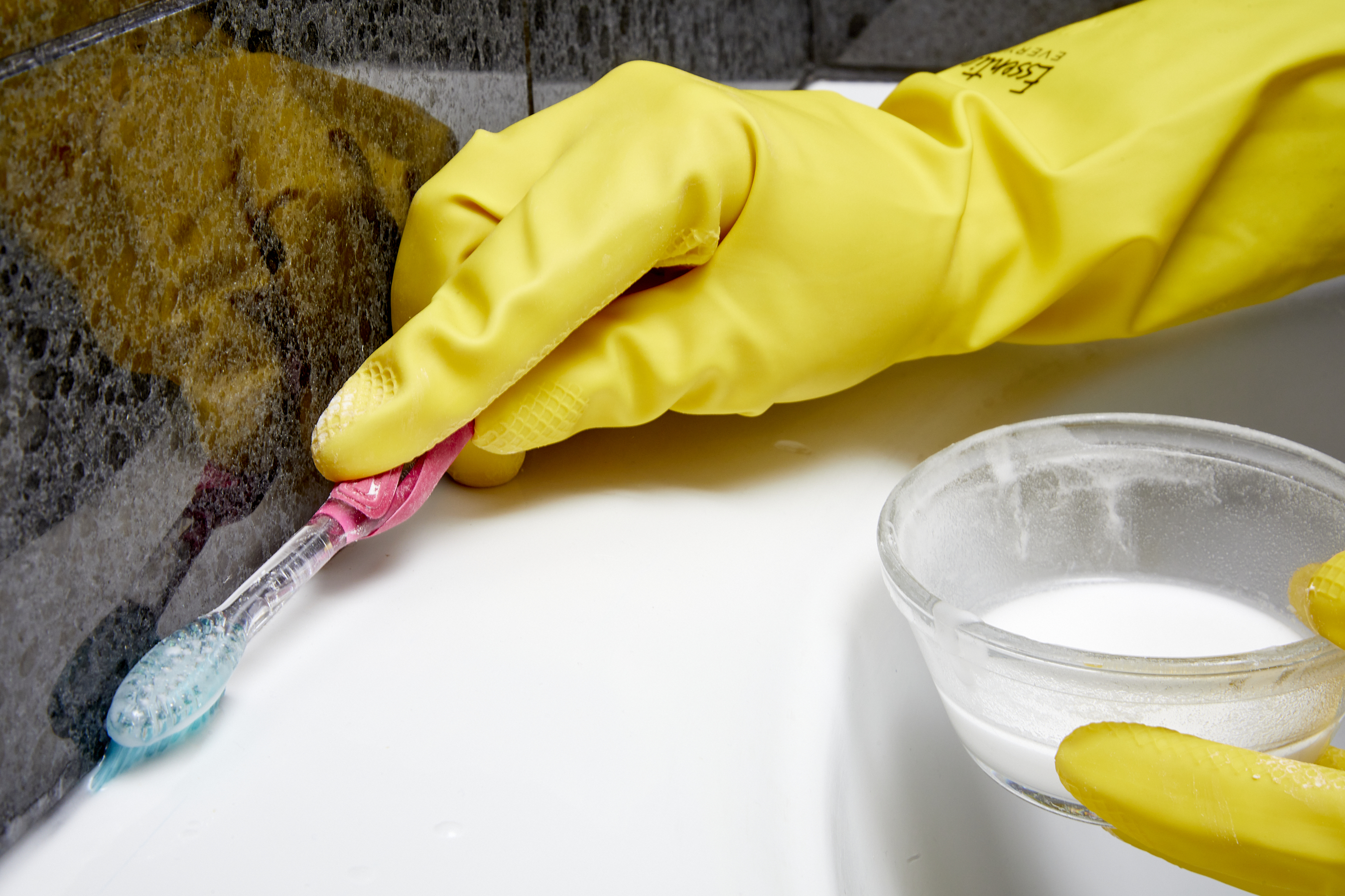 Person wearing rubber gloves uses toothbrush to clean crevices around bathtub.