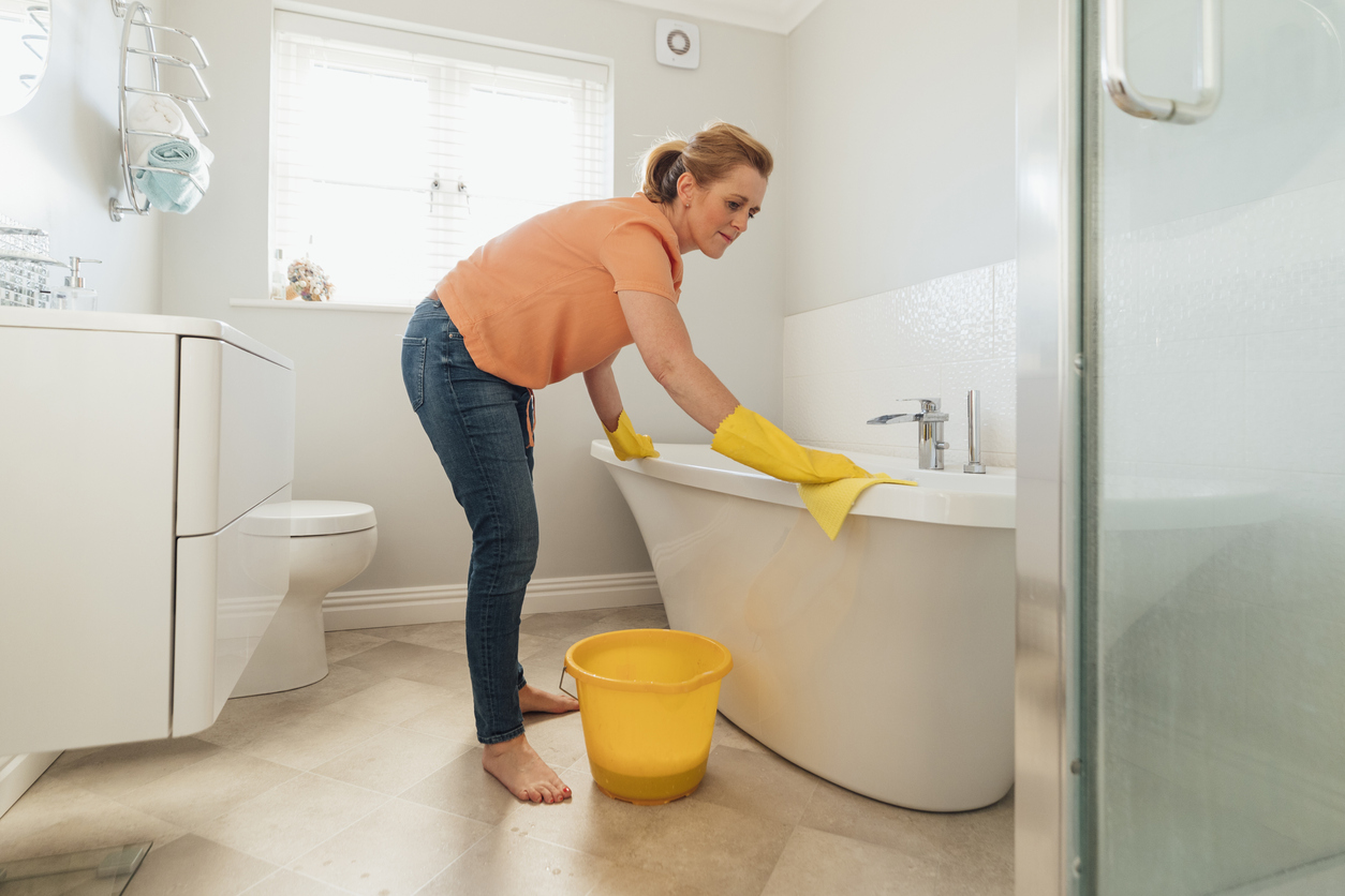 A mid-adult caucasian woman wearing protective yellow gloves is cleaning the bathtub with soap and water in her bathroom at her home in Hartlepool.