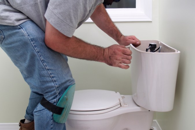 A man is inspecting a toilet with the tank lid removed with the intention of repairing it.