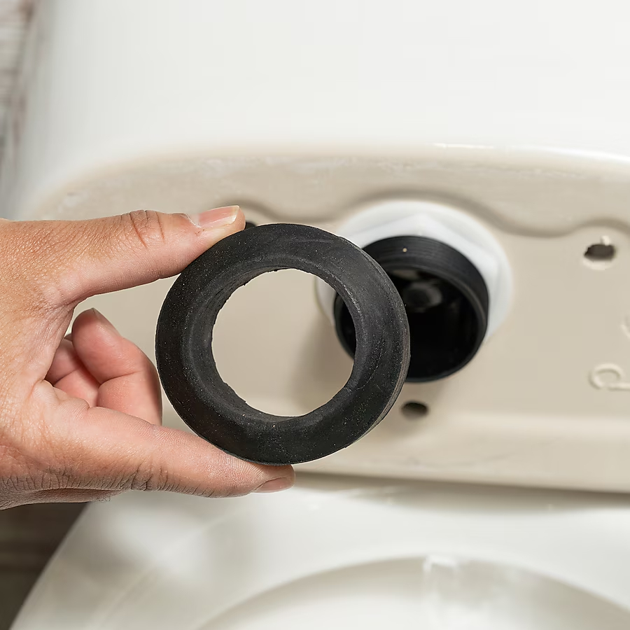 A person holds a black toilet tank-to-bowl gasket in front of a toilet tank ahead of installation.