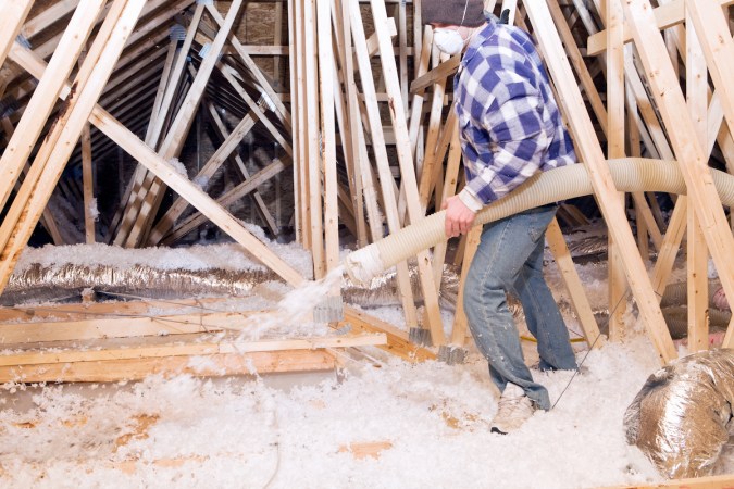 Worker sprays blown in fiberglass insulation in attic.