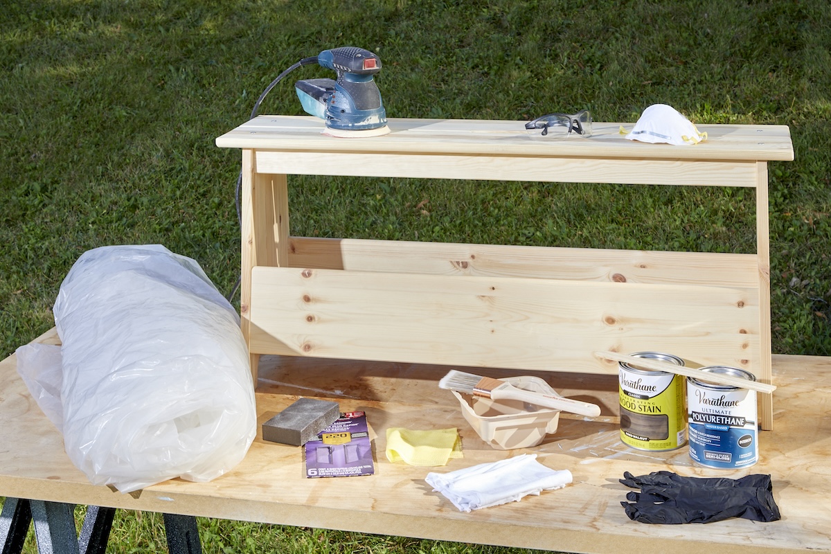 Gathering the materials needed to stain a wooden shelf like wood stain, rubber gloves, brushes, a plastic sheet, mask, and sander.