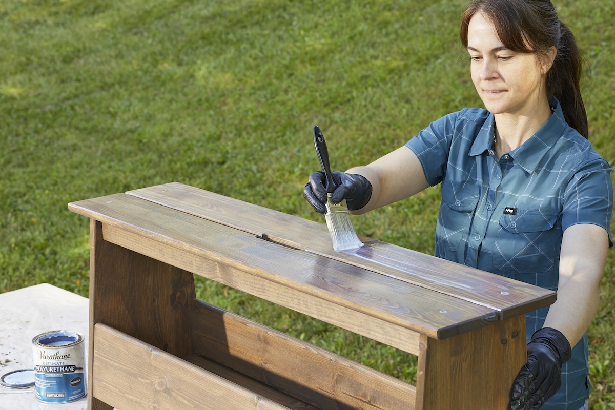 Person in blue blouse applies a wood sealant onto a freshly stain wooden shelf with a paint brush.