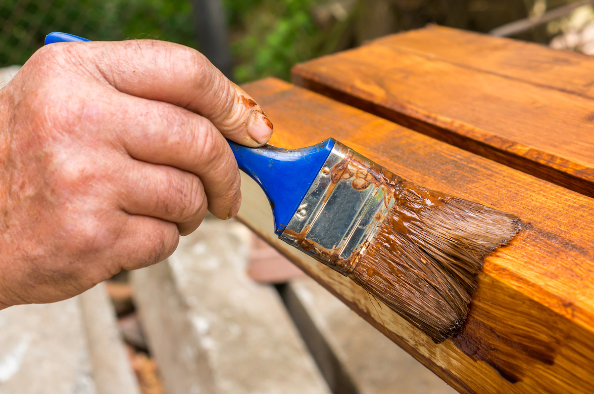 Painter uses a paintbrush to stain the wooden slats of an outdoor chair.