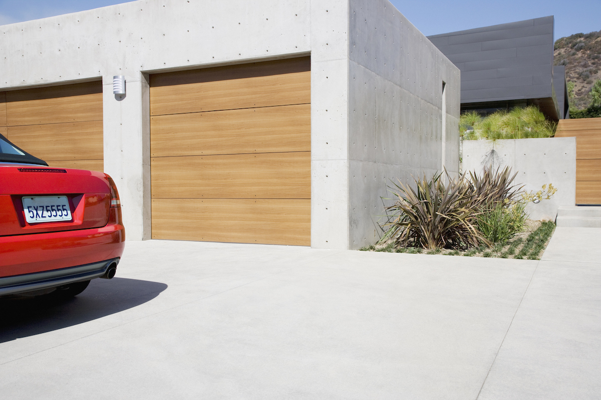 A red car is parked outside of a concrete two-car garage with wooden doors.