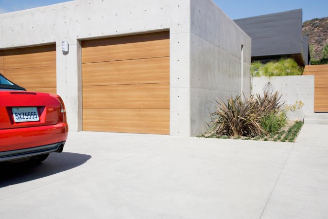 A red car is parked outside of a concrete two-car garage with wooden doors.