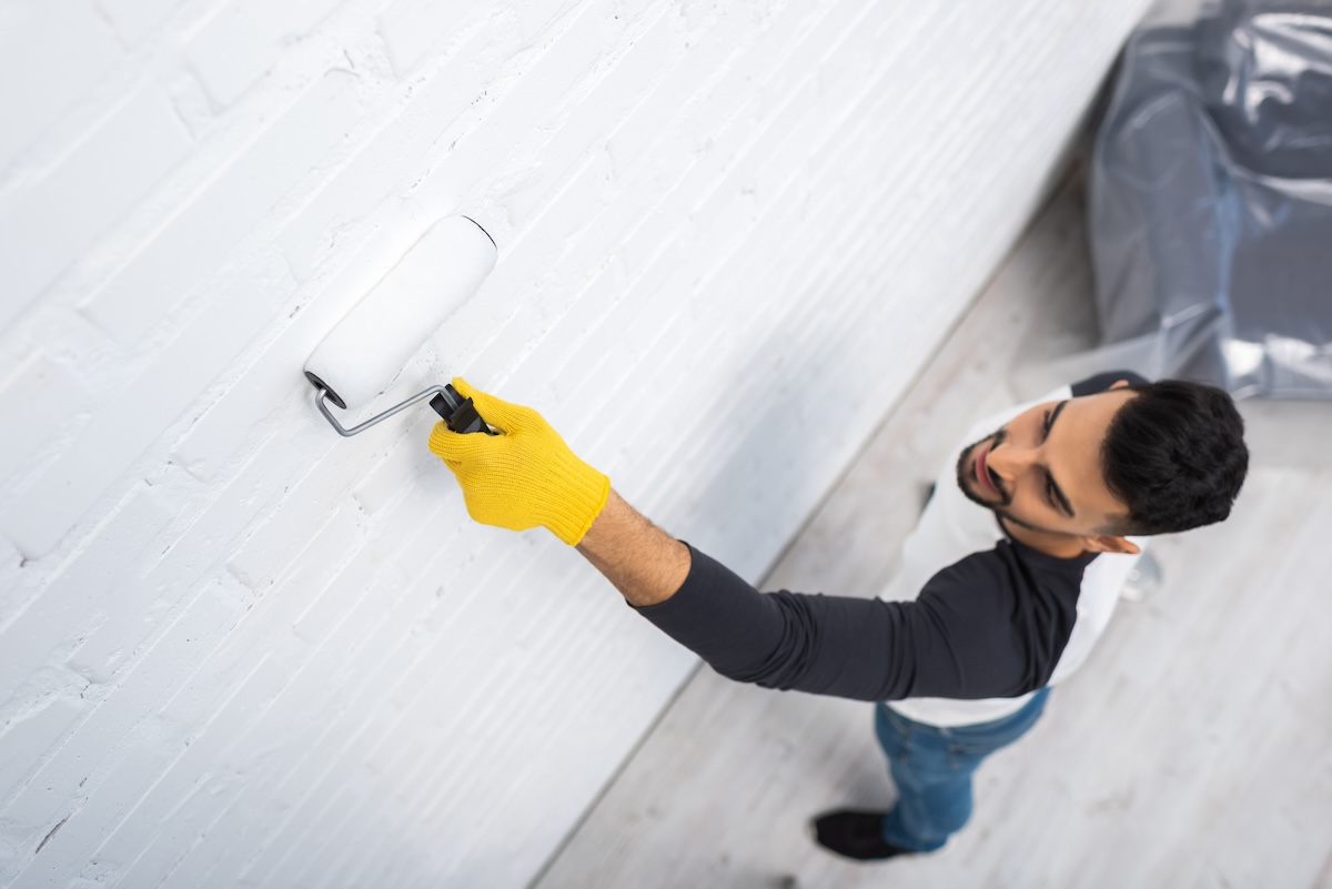 A man wearing a pair of yellow gloves paints a second coat of white paint on a brick wall. 