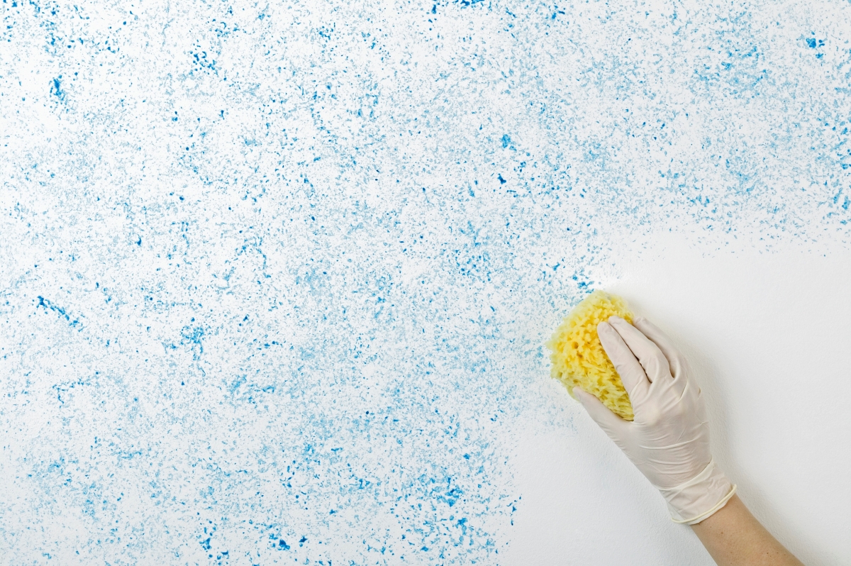 A gloved hand is using a yellow sponge to add blue colored texture to a white wall.