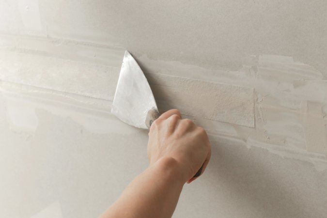 Person uses a putty knife to skim coat over drywall tape.