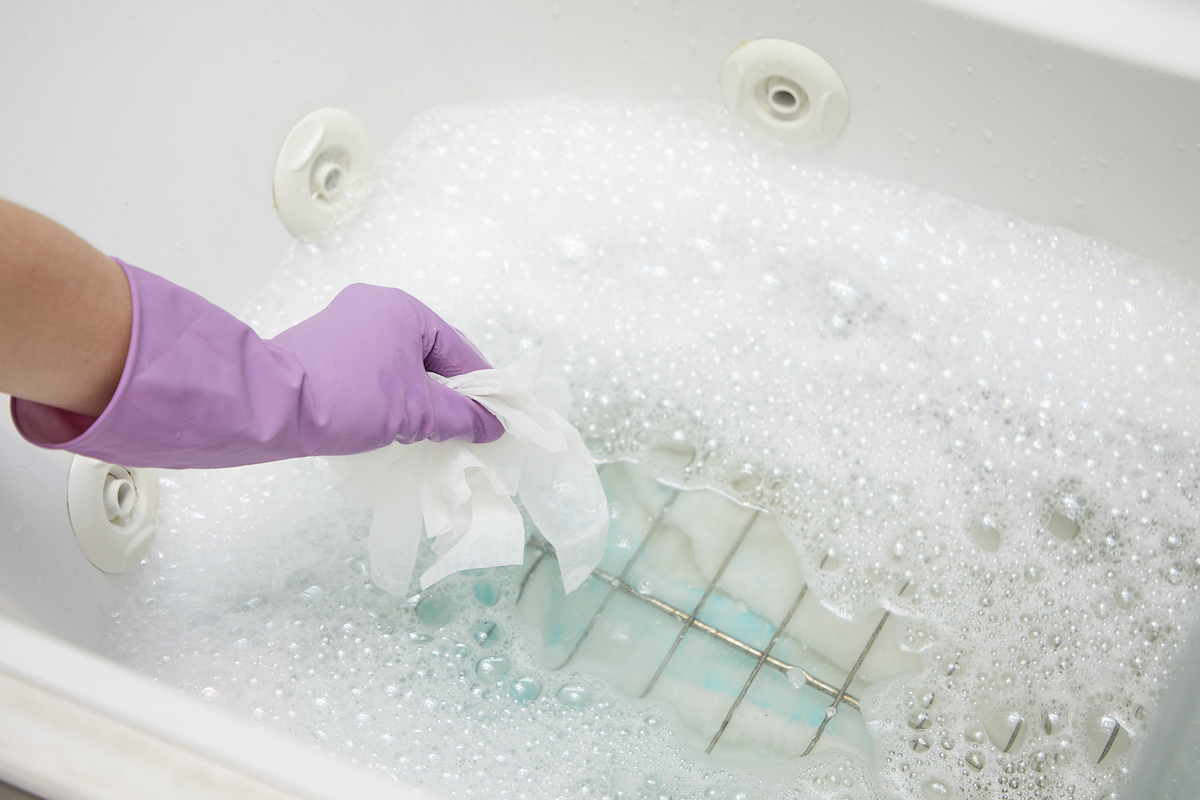 Woman wearing rubber gloves uses dryer sheets and dish soap to clean oven racks in a bathtub.