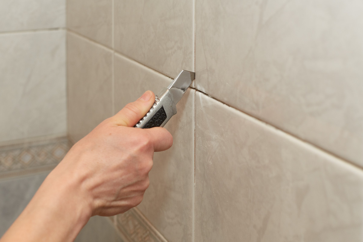 Male hands with knife removing the old grout from beige tile.