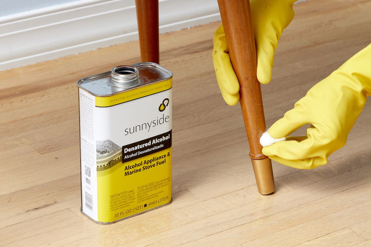 Woman rubs a cotton ball onto the leg of a wood table, a container of denatured alcohol nearby.