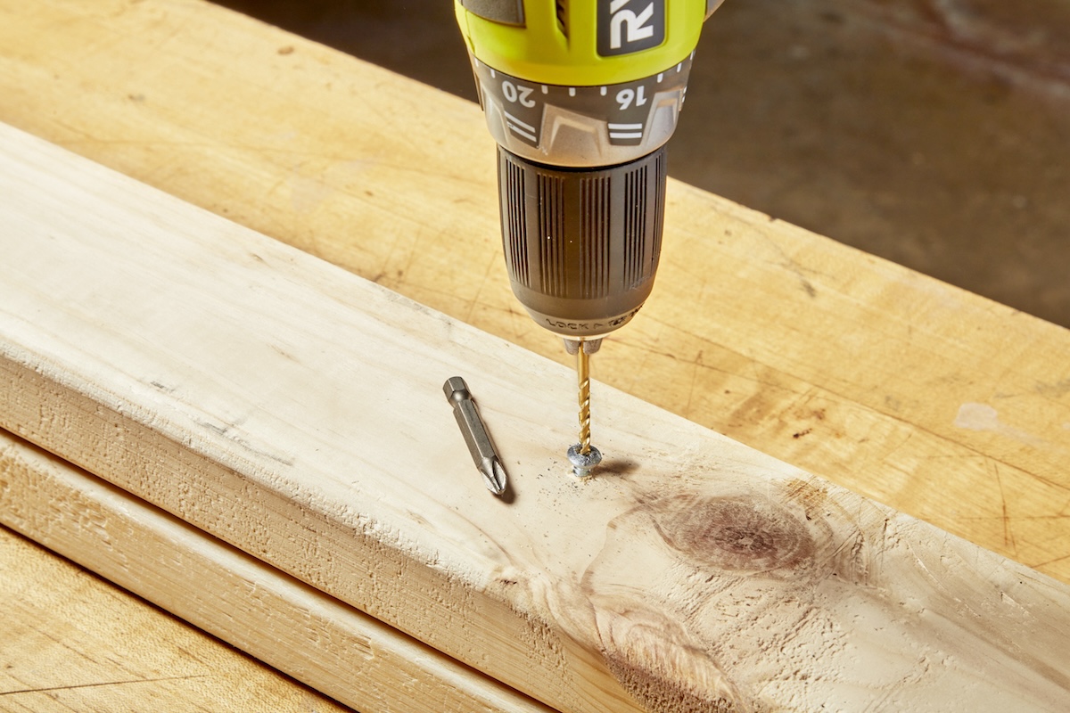 A person using a metal drill bit to drill into the head of a stripped screw stuck in a wood stud.