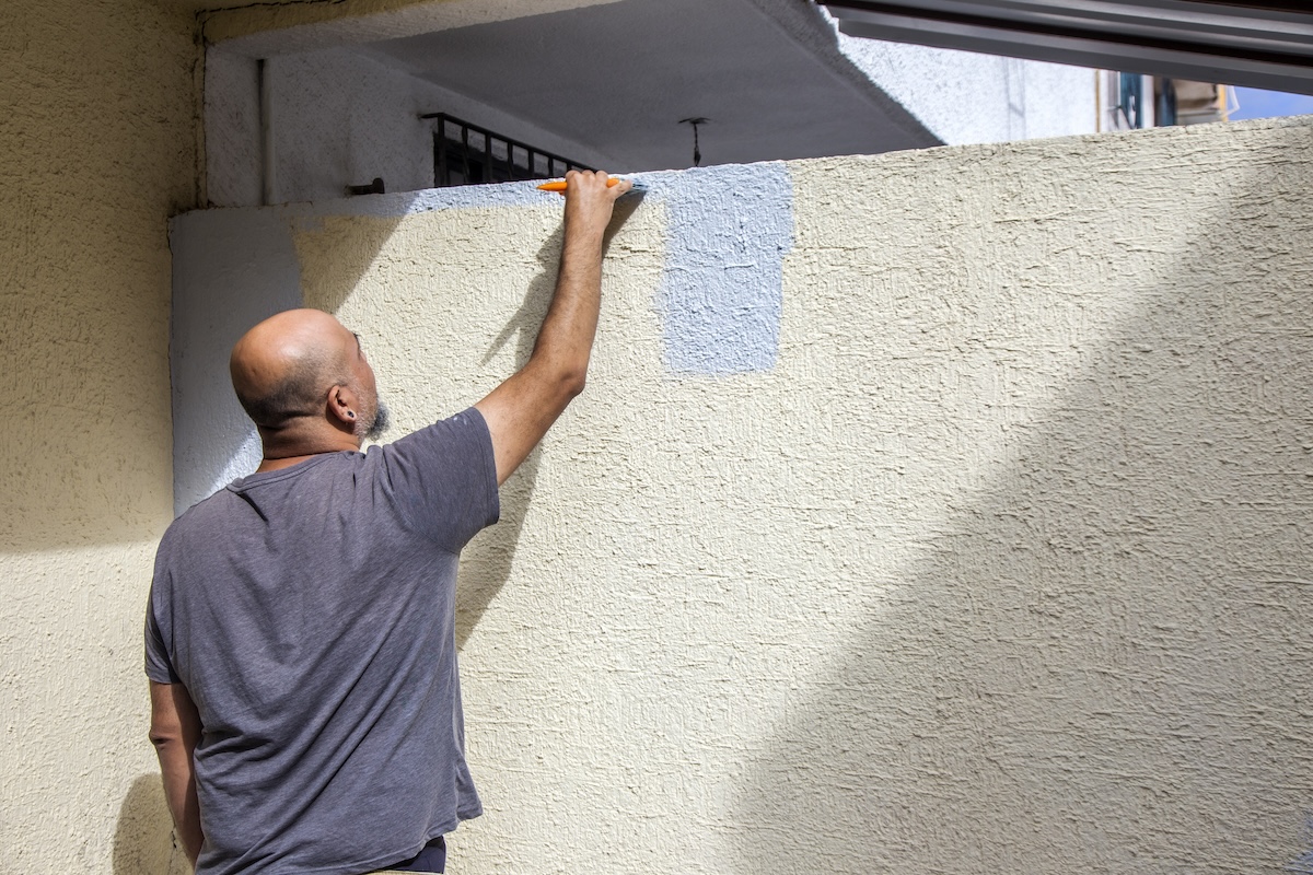 A man in a gray shirt paints an exterior concrete wall with yellow paint.