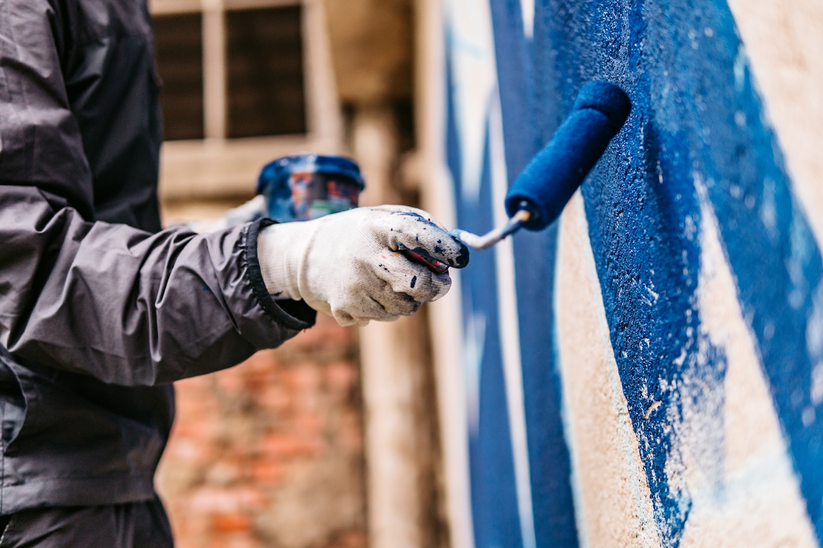 A worker with white gloves uses a roller brush to paint a concrete wall blue.