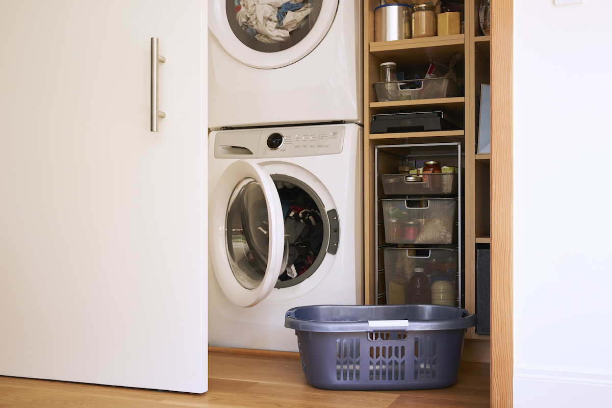 A blue laundry basket on the floor is in front of a stackable washing machine and dryer, which are next to pantry items.