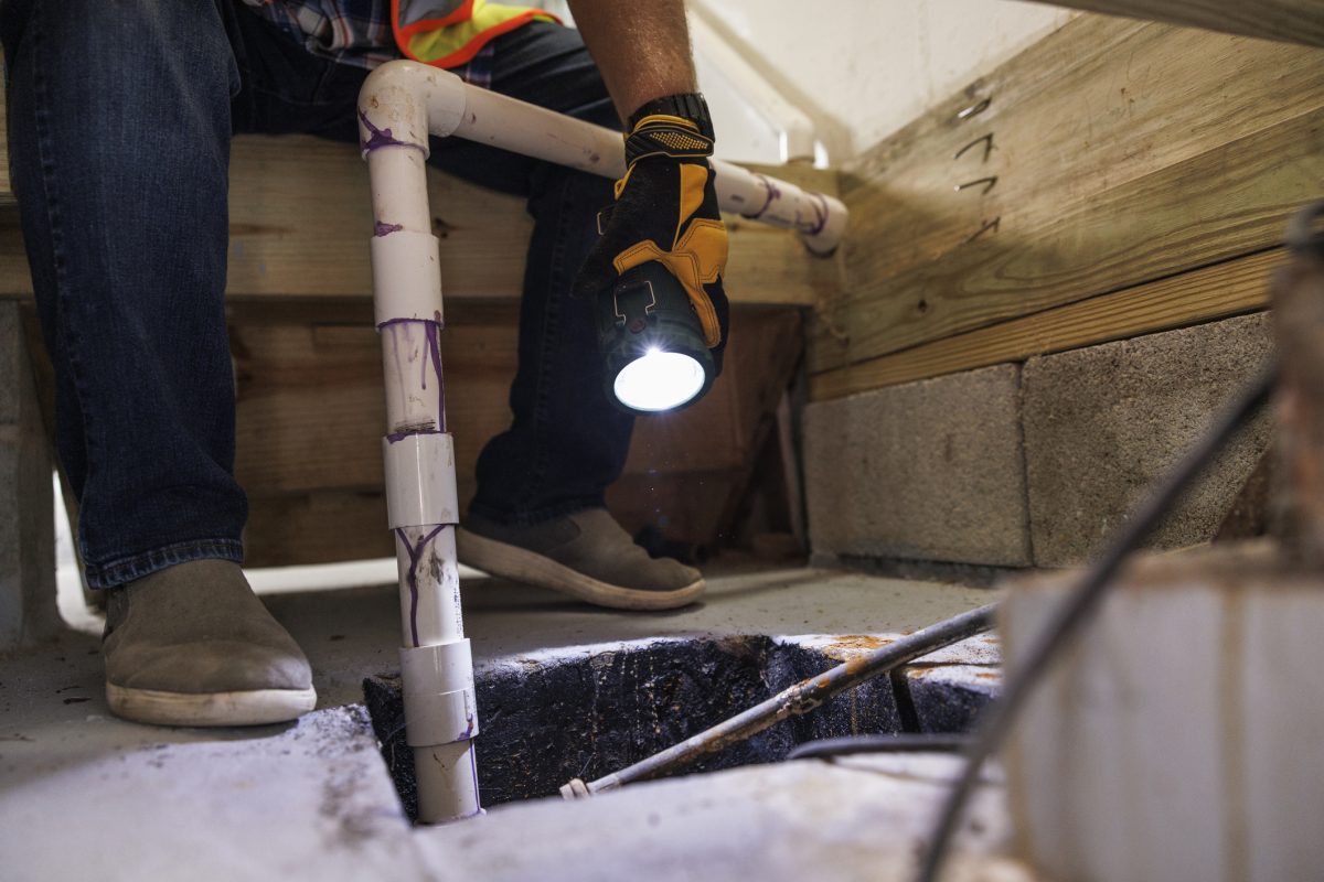 A gloved man points a flashlight into the sump pit of a residential basement.