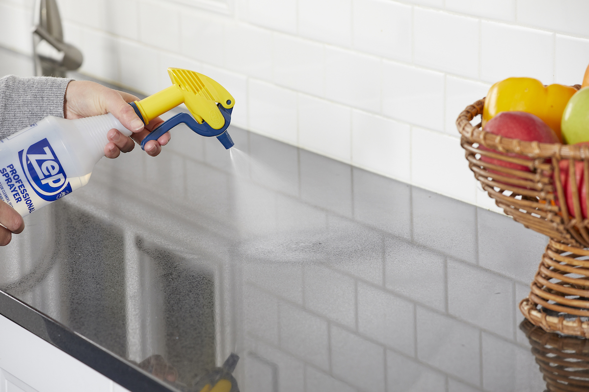 Woman points spray bottle at a grey kitchen countertop, a bowl of fruit nearby.