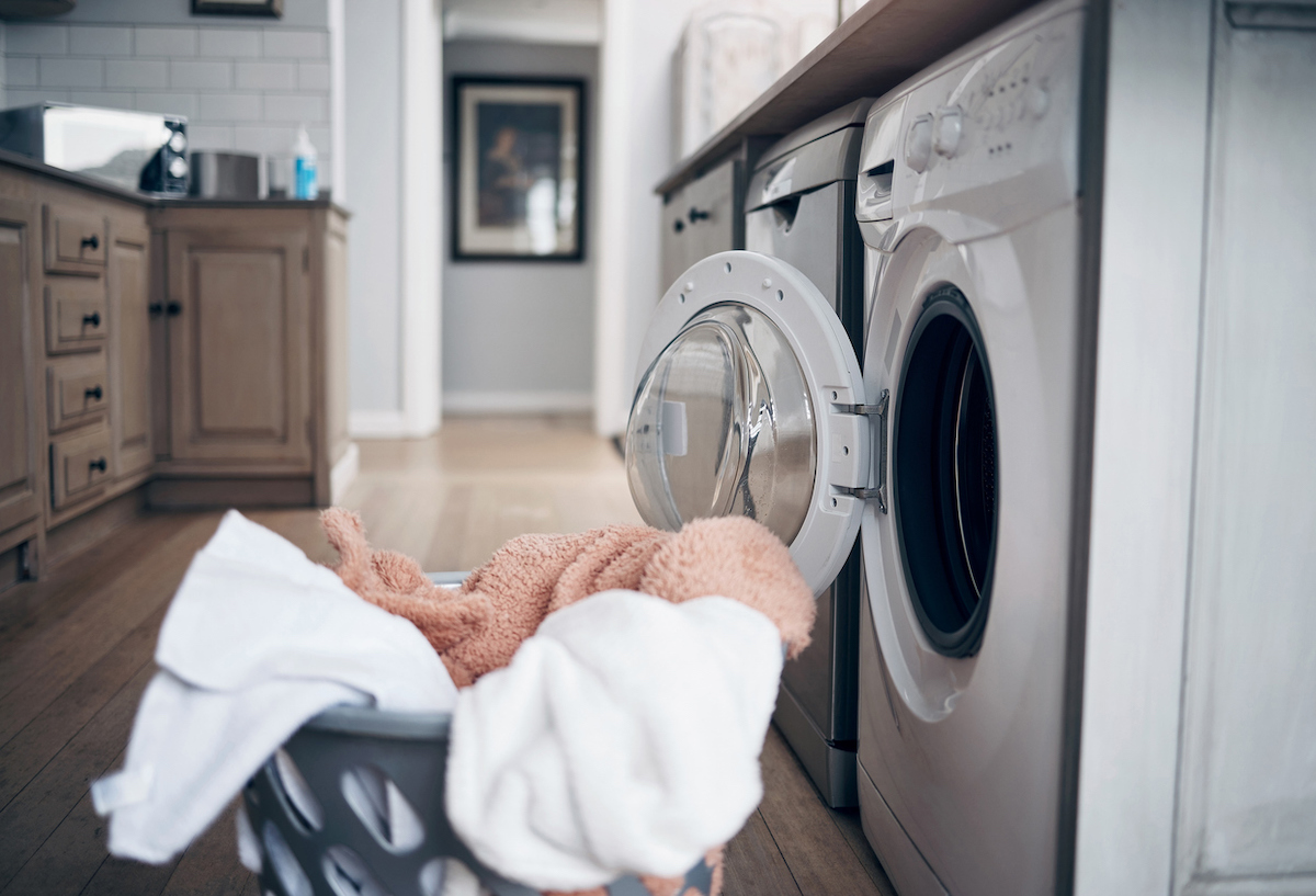 A blue laundry basket full of clothes is on the floor in front of an open washing machine that is underneath a kitchen counter and next to a dishwasher.