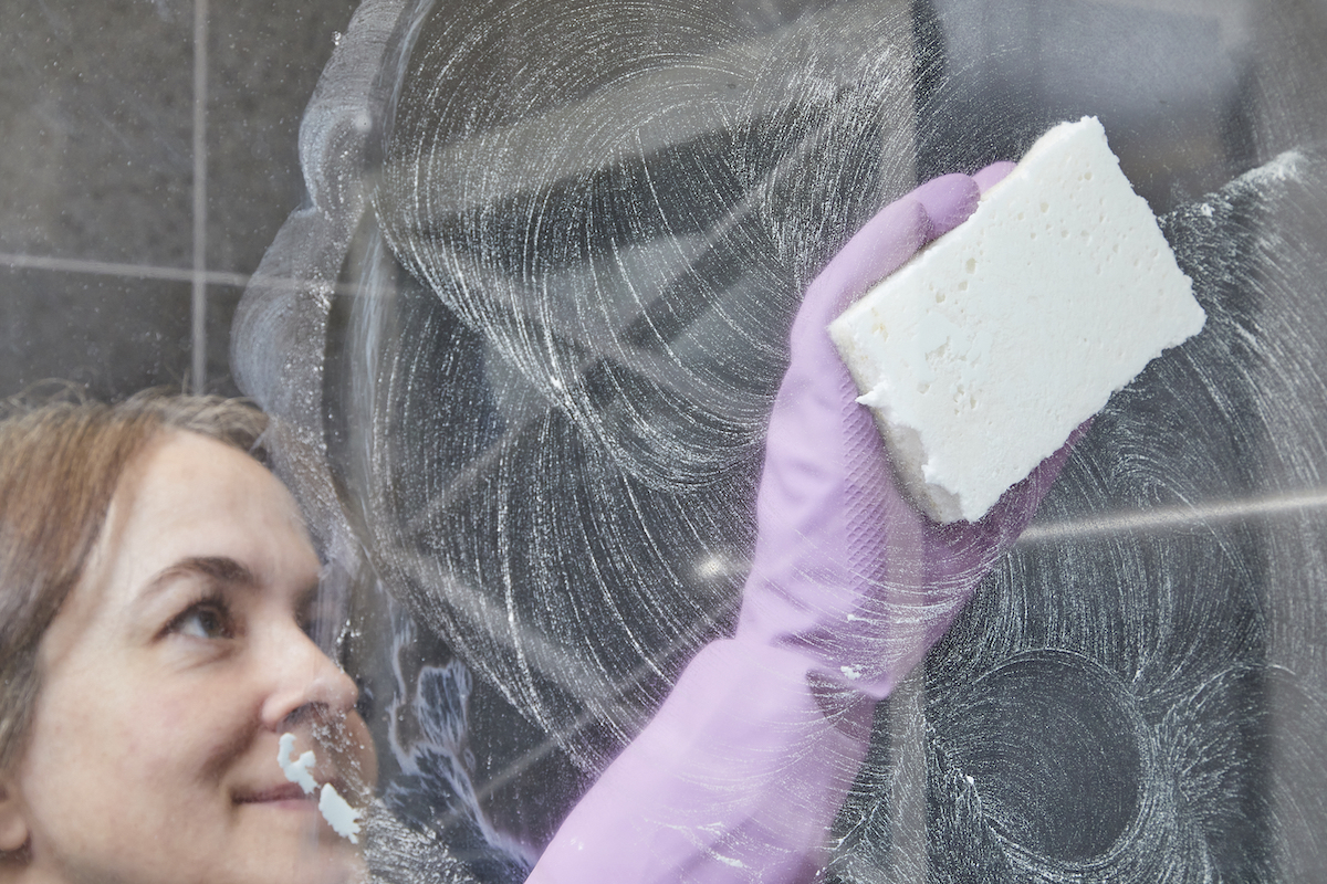 Woman in rubber gloves cleans a shower door with a sponge.