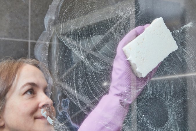 Woman in rubber gloves cleans a shower door with a sponge.