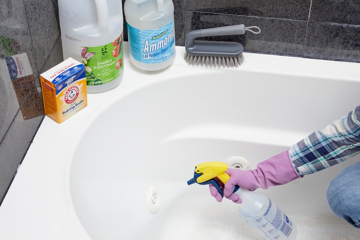 Woman points spray bottle at her bathtub; containers of baking soda, vinegar, ammonia, and a scrub brush on ledge nearby.