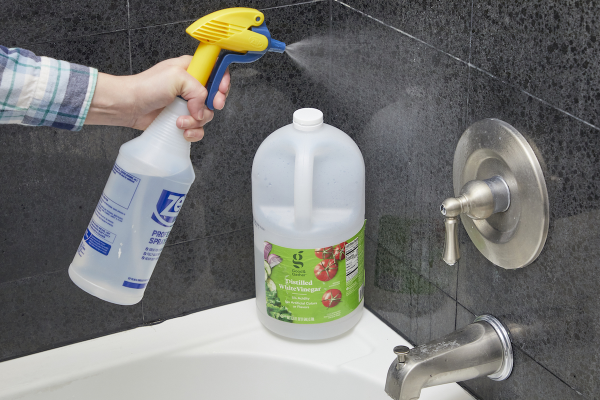 Woman points a spray bottle at a bathtub wall; large bottle of vinegar on the ledge in the background.