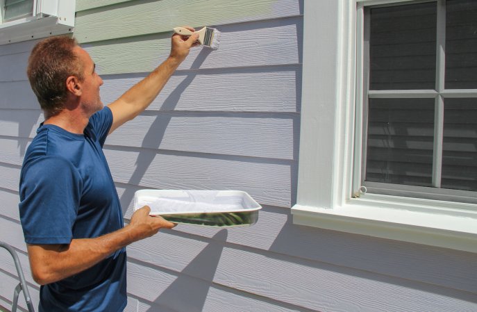 Man holding a paint tray in right hand and painting the gray exterior siding of a home with a paintbrush in his left hand.