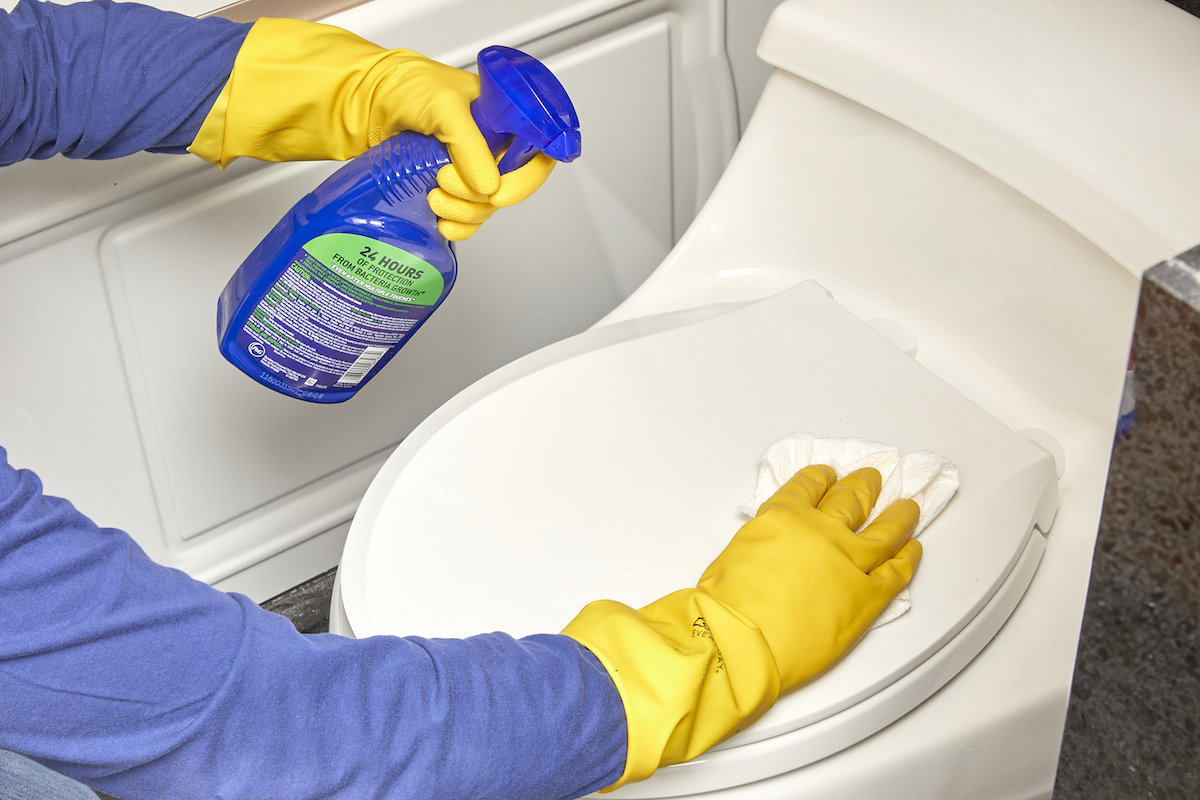 A woman wearing rubber gloves using spray cleaner to clean the outside of a toilet.