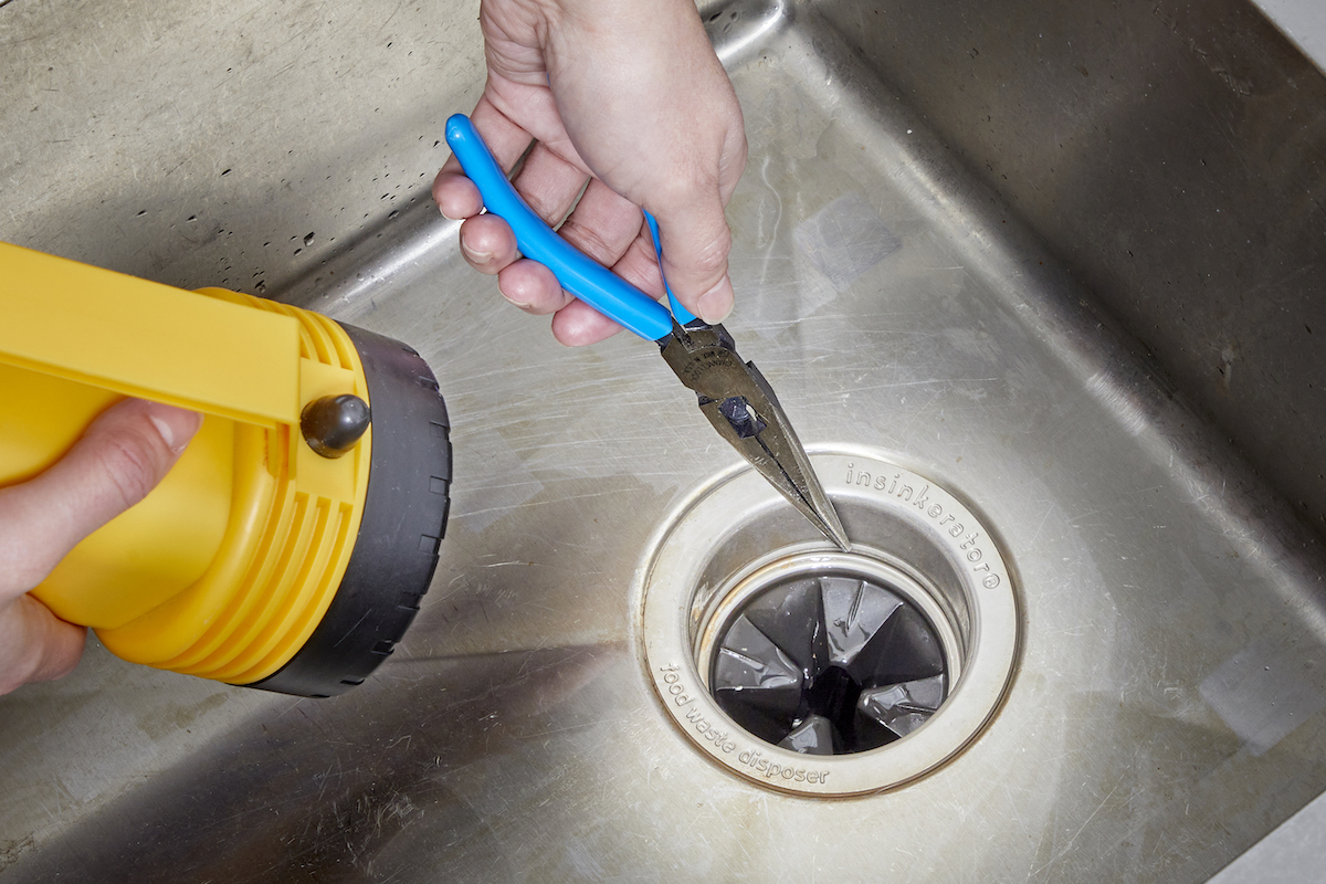 Woman points needle-nose pliers toward garbage disposal, large flashlight in the other hand.