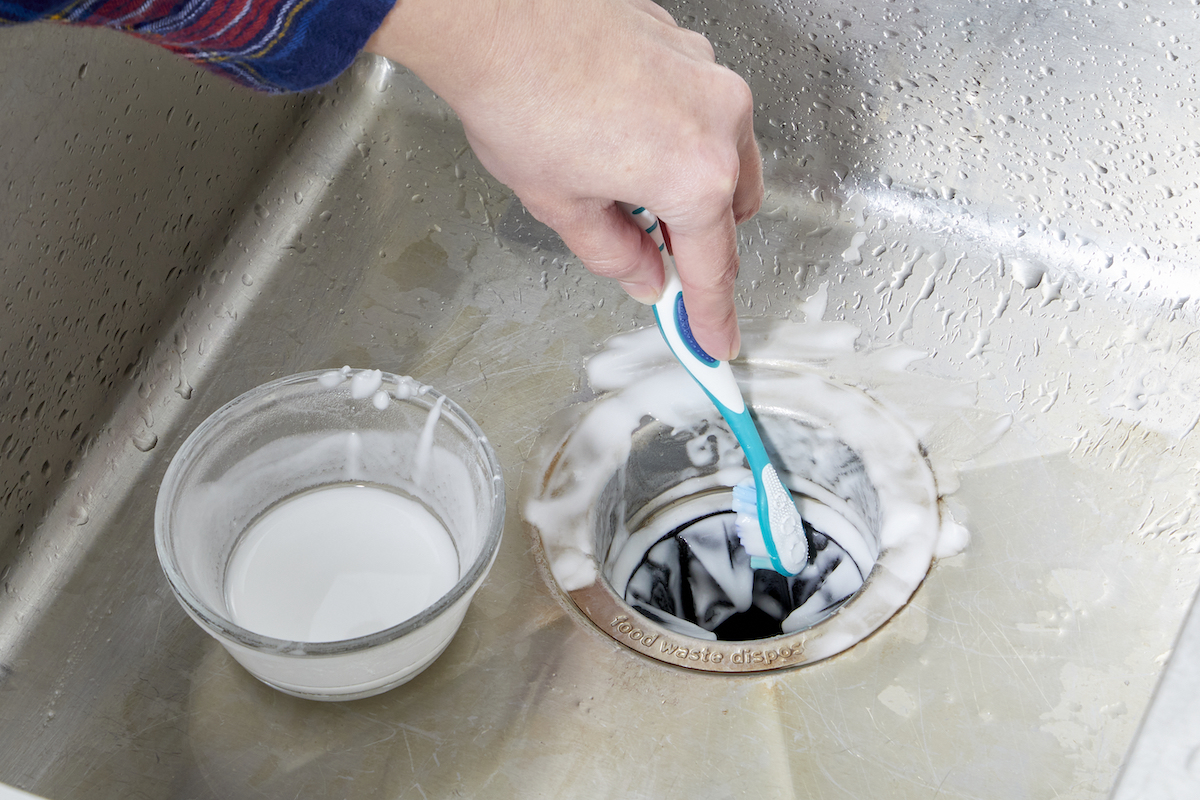 Woman scrubs the rubber flaps inside her garbage disposal with a baking soda and vinegar paste, in a bowl beside the disposal.