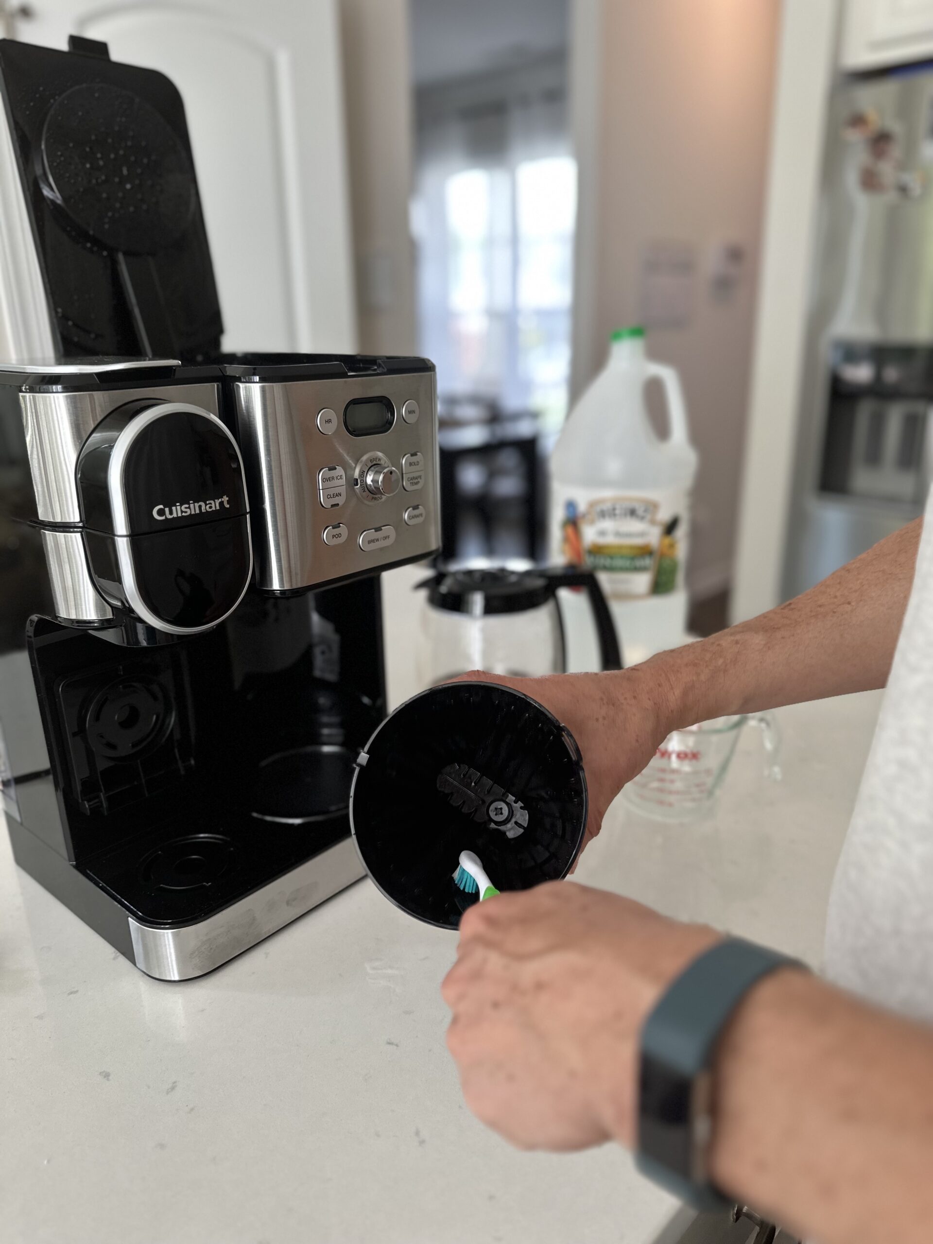 using a toothbrush to clean out basket of coffee maker with coffee maker sitting on kitchen counter