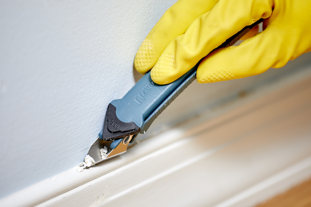 Woman wearing yellow gloves uses a caulk remover tool to remove caulk from atop a baseboard.