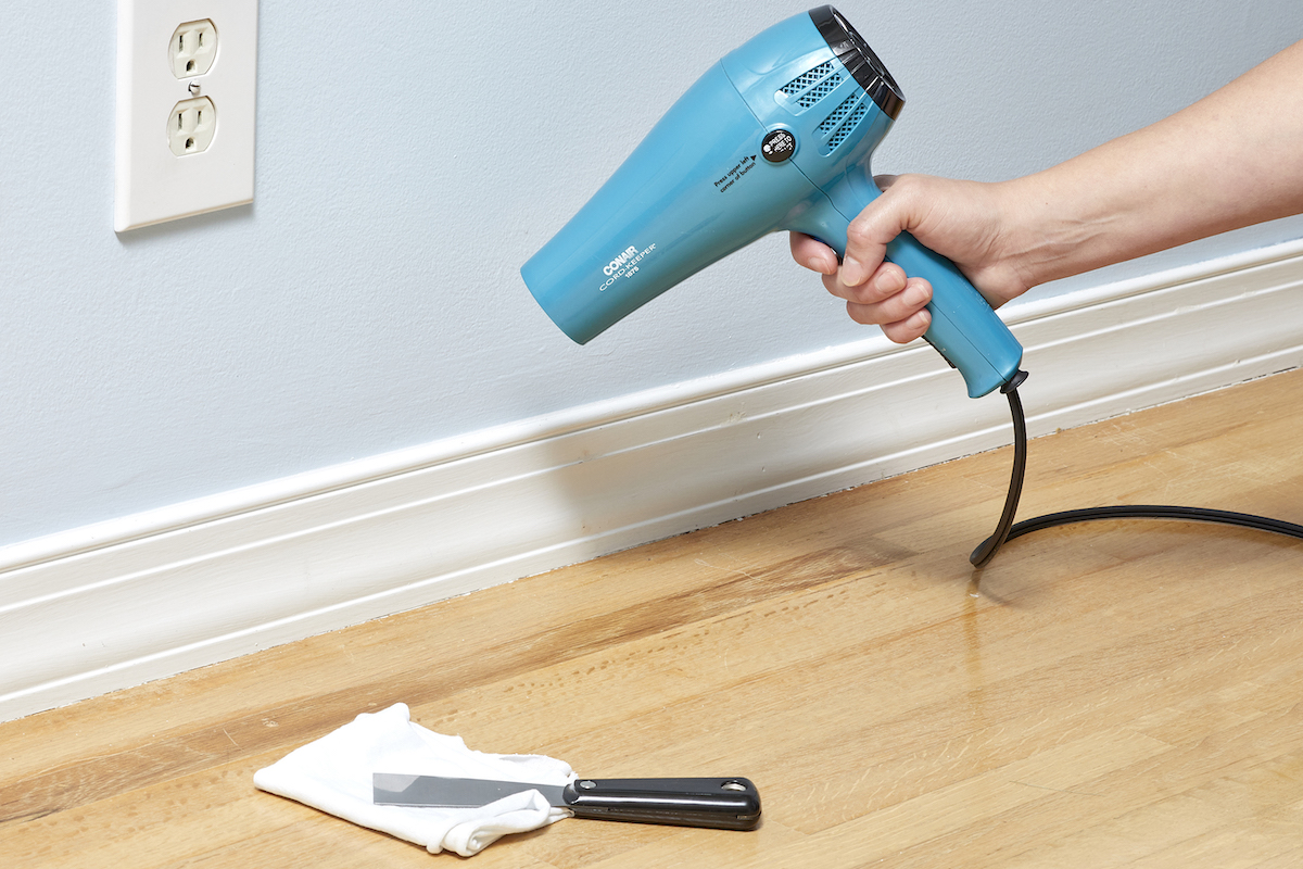 Woman points hair dryer at caulk along a baseboard wall.