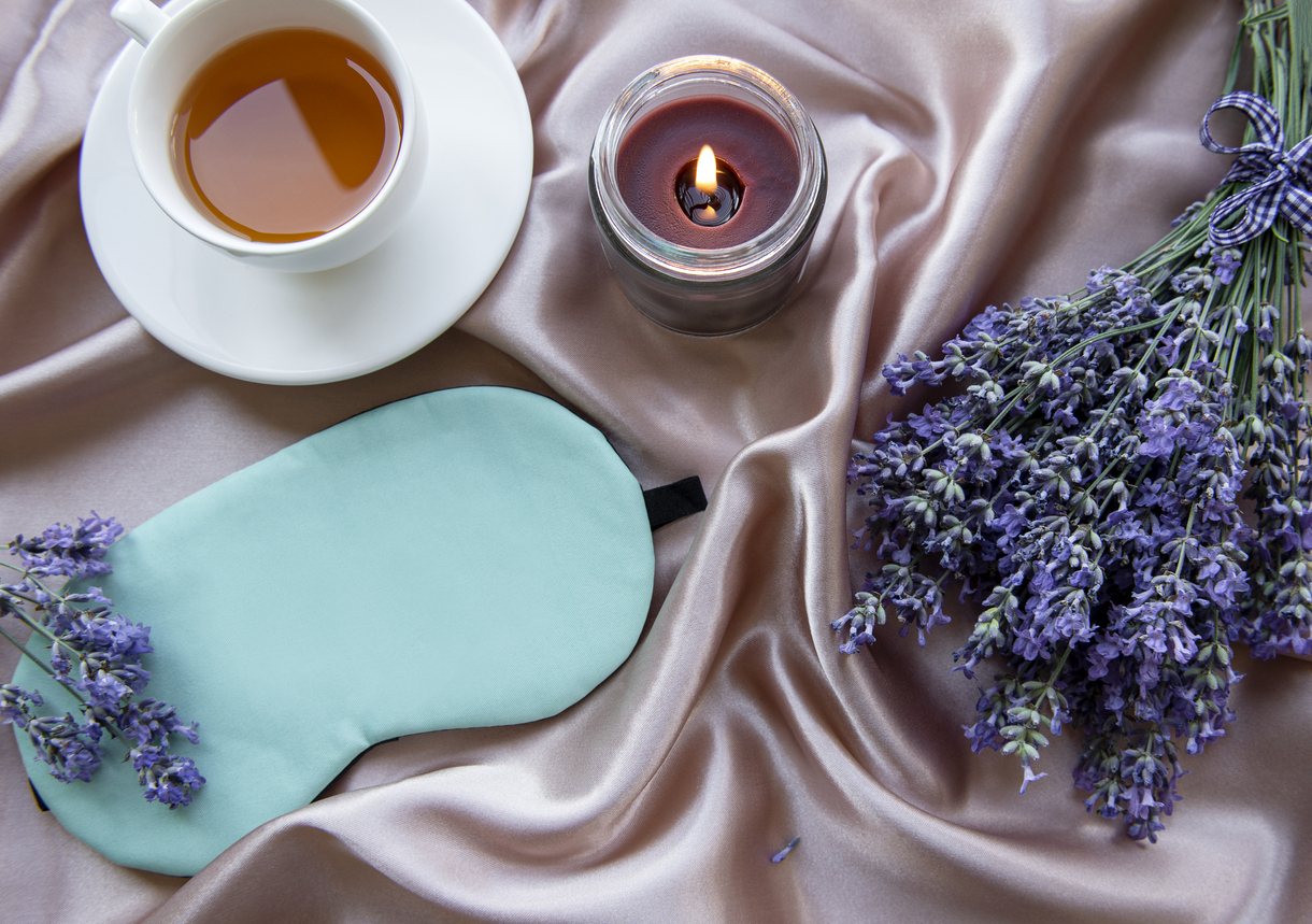 overhead view of lavendar plant laid out on pink silk with sleep mask and lavender tea and a lit lavendar candle