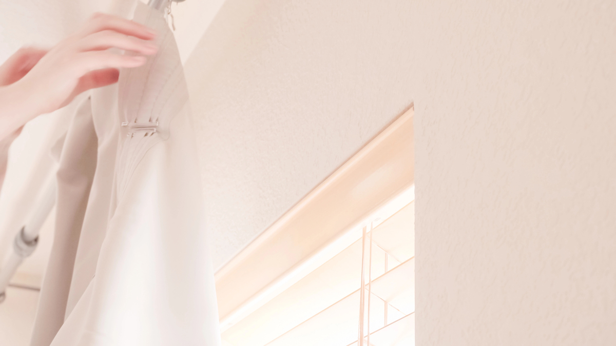 Woman hangs curtains on a curtain rod in a room with light pink walls.