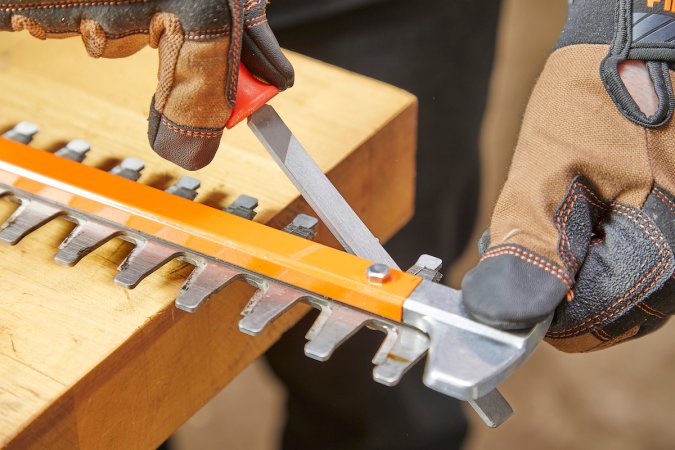 Sharpening an orange hedge trimmer on work bench with work gloves on.