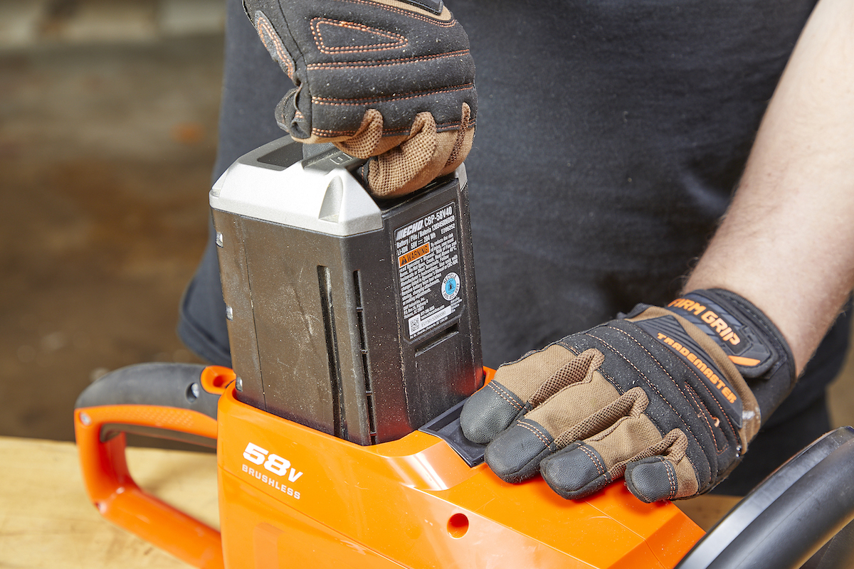 Unplugging the battery from an orange hedge trimmer with work gloves on.