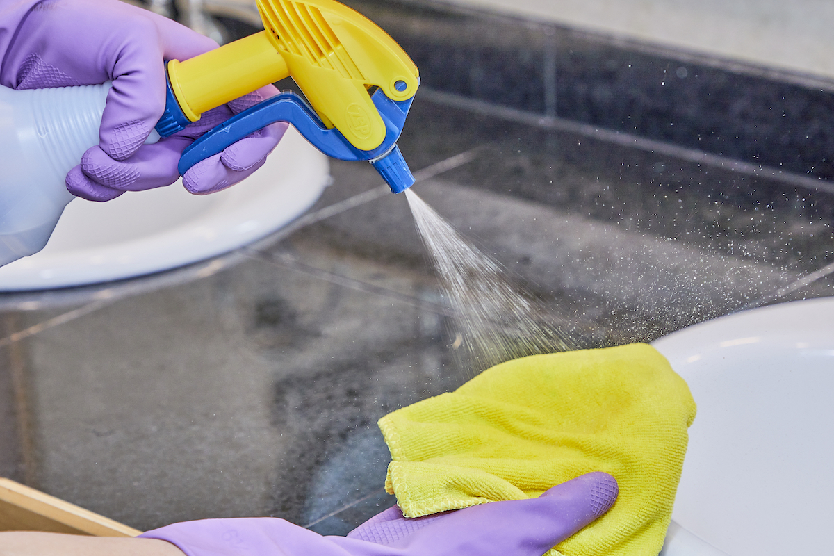 Woman wearing purple cleaning gloves sprays a yellow microfiber cloth with cleaning solution.