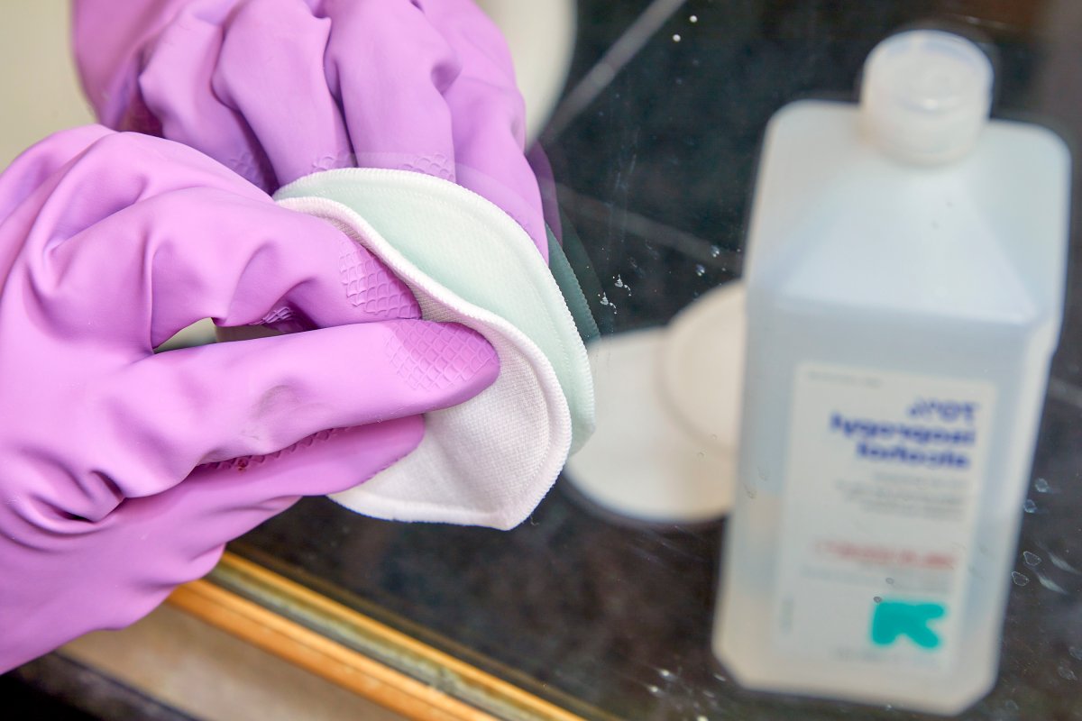 Woman wearing pink cleaning gloves uses rubbing alcohol and a cotton pad to remove residue from a mirror.