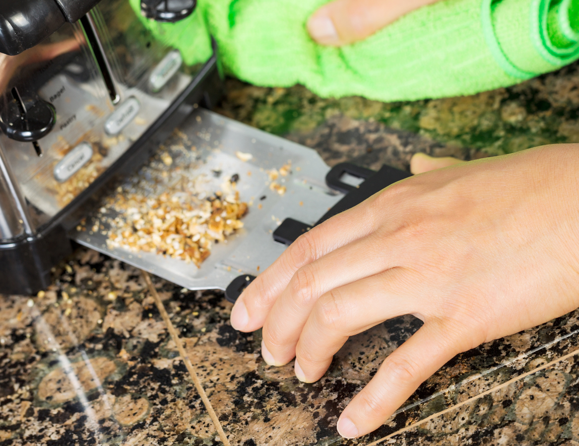 Photo of female hand cleaning bottom of kitchen toaster tray with green microfiber rag with stone counter top in background